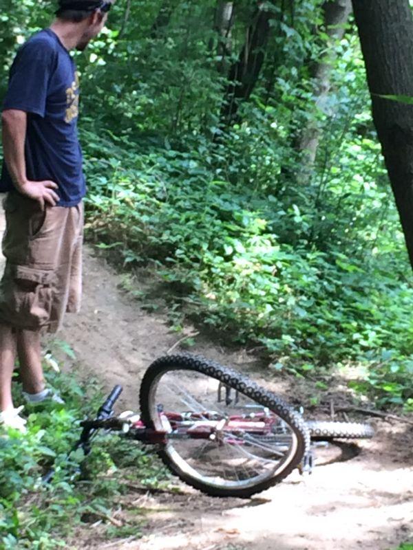 A person in a blue shirt and cargo shorts stands next to a fallen mountain bike on a dirt trail surrounded by greenery. The bike is on its side, partially off the trail, with the wheels visible and vegetation in the background. Rolling Hills Park mountain bike trail.
