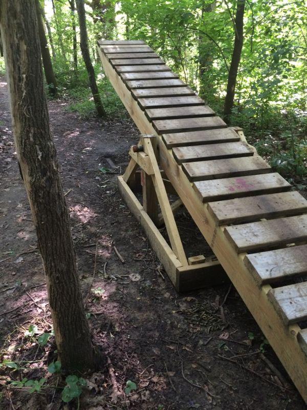 Wooden ramp constructed on the edge of a forest path, surrounded by trees and greenery. The ramp is angled and has a slatted surface, designed for outdoor activities. Memorial Park mountain bike trail.