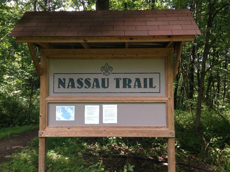 Signpost for Nassau Trail, featuring a wooden structure with a sloped roof and a large, bold title. Below the title, there are informational panels, set against a backdrop of lush greenery and trees. Nassau Trails mountain bike trail.