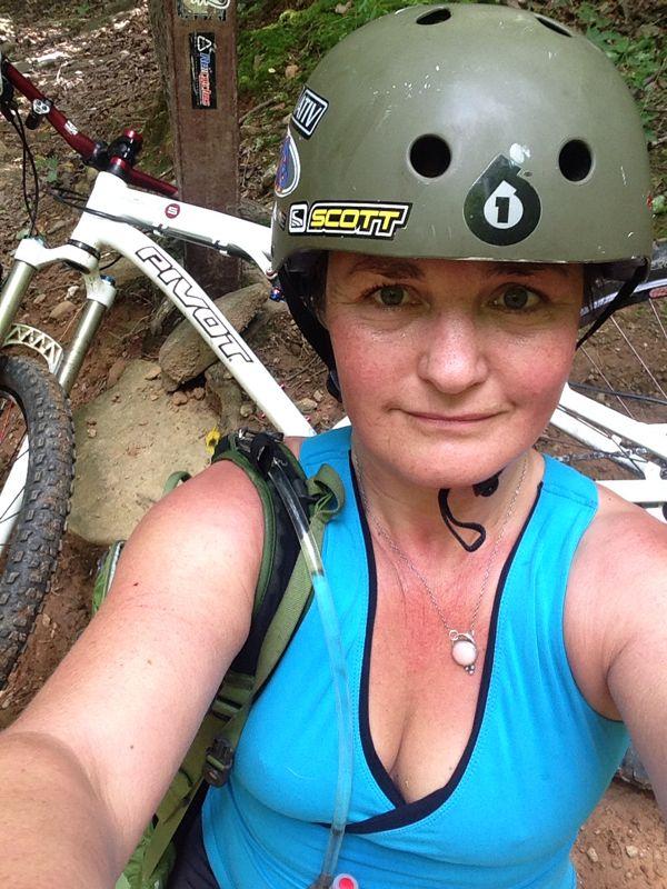 A woman wearing a green helmet and a blue tank top poses for a selfie while resting next to her mountain bike on a wooded trail. She has a backpack on and is smiling, with the bike leaning against a tree in the background. Bent Creek mountain bike trail.