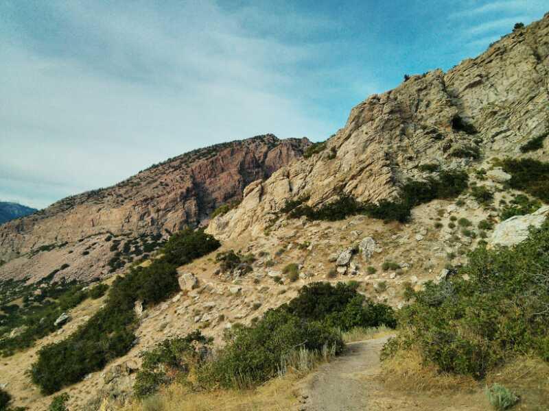 A rocky landscape featuring a winding dirt path leading up a hillside, surrounded by sparse vegetation and rocky outcrops against a backdrop of blue skies with scattered clouds. Bonneville Shoreline Ogden South Of 12th mountain bike trail.