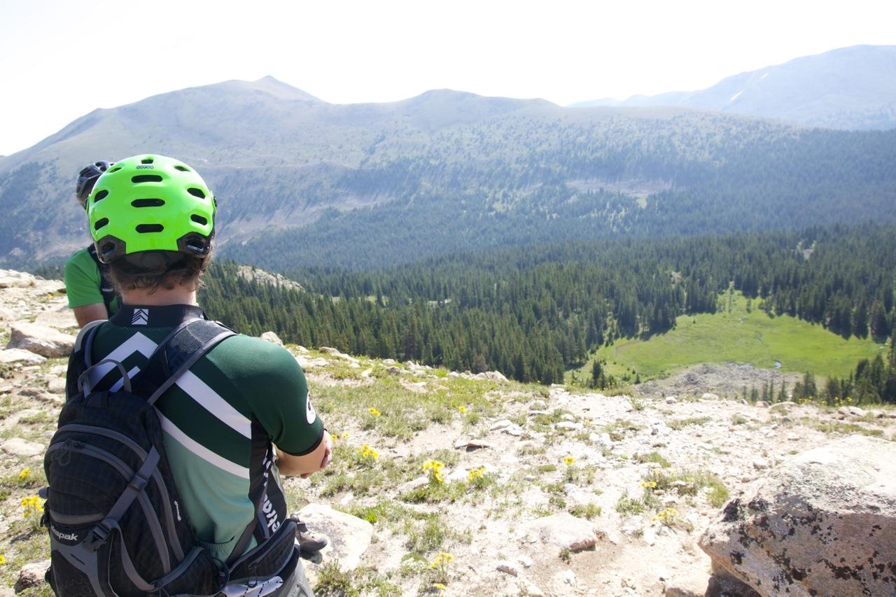 A person wearing a bright green helmet and a sporty shirt stands on a rocky ledge, gazing out over a vast landscape of mountains, forests, and meadows. The scene captures a beautiful day in the wilderness, showcasing greenery and distant peaks under a clear sky. Monarch Crest Trail mountain bike trail.