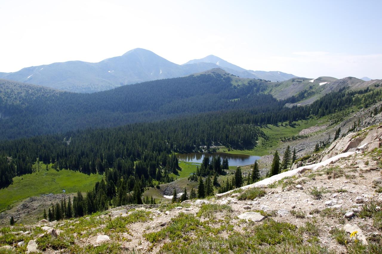 A panoramic view of a mountainous landscape featuring lush green forests and a tranquil lake nestled in a valley. The scene shows rolling hills in the background, with several peaks reflecting distant snow. The foreground includes rocky terrain with scattered wildflowers. Monarch Crest Trail mountain bike trail.