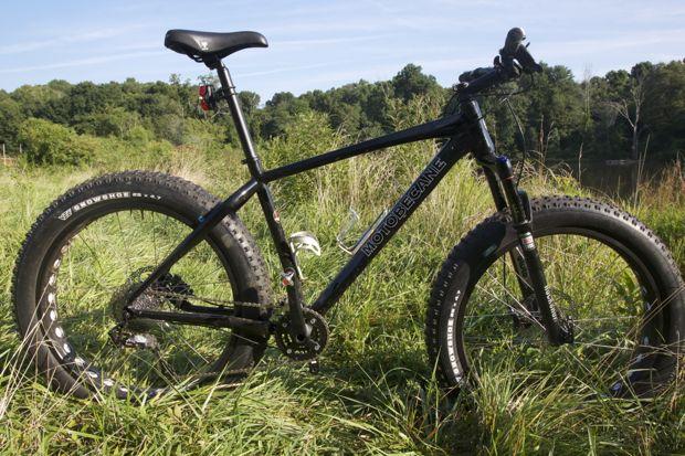 Motobecane NightTrain Bullet: A black fat tire mountain bike is positioned on a grassy area, with a backdrop of trees and a clear sky. The bike features wide tires designed for off-road terrains and a sturdy frame, showcasing its capability for rugged outdoor adventures.