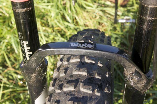 Motobecane NightTrain Bullet: A close-up view of a bicycle front fork with "Bluto" branding, positioned over a thick, knobby tire. The background features green grass, suggesting an outdoor setting.