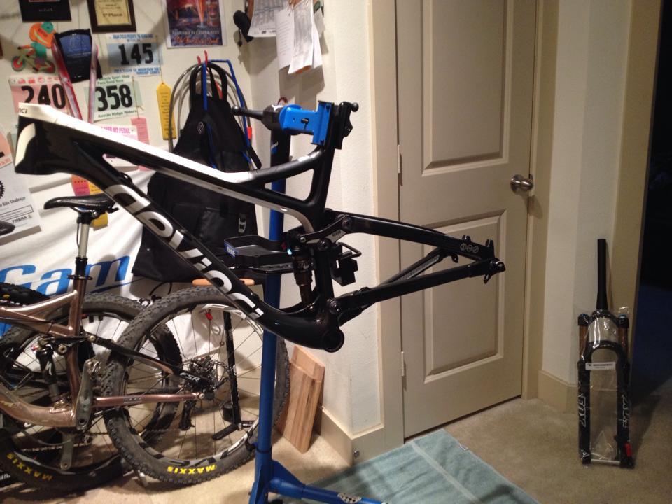 Devinci Atlas Carbon SL: Image of a bicycle frame mounted on a repair stand, with various biking accessories and tools visible in the background. There are also two mountain bikes leaning against the wall, along with several race number bibs and awards pinned up. A door is visible in the background, and a bicycle fork is propped up against the wall.