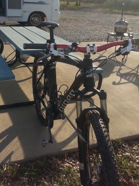 Niner EMD 9: A close-up view of a mountain bike parked on a concrete surface, showcasing its handlebars, front suspension, and tires. In the background, there is a picnic table and a campsite with an RV and a grill, surrounded by natural scenery.