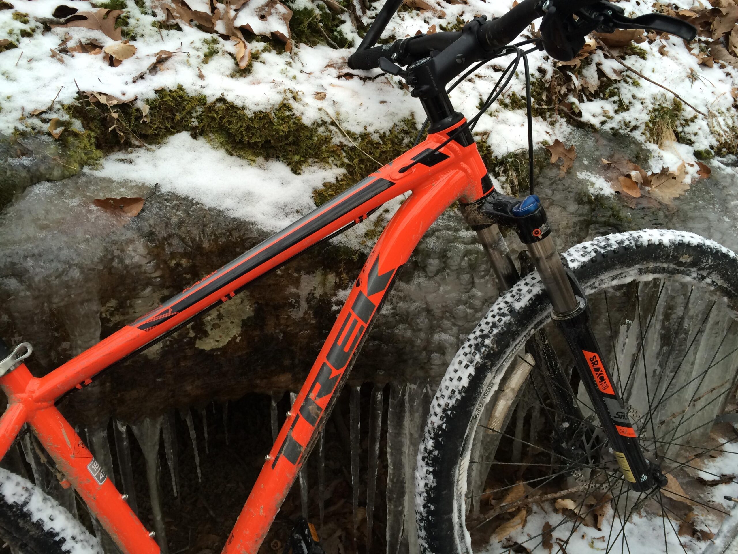 Trek X-Caliber 7: Close-up of an orange Trek mountain bike positioned near a snowy, icy landscape with fallen leaves in the foreground. The bike features a sturdy frame and wide, textured tires. Icicles hang from a rock in the background, highlighting the cold environment.