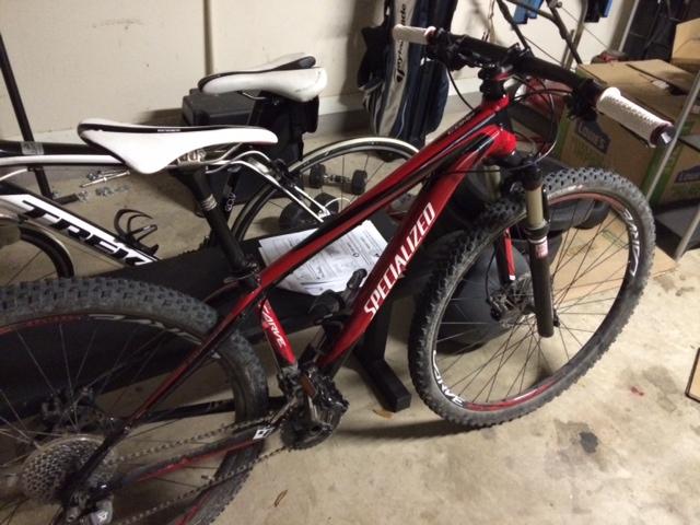 Specialized Carve 2013 comp: A red Specialized mountain bike positioned in a garage, with another bike partially visible in the background. The bike features thick tires and a front suspension fork. Papers and a black bag can be seen on the floor nearby, indicating a space used for storage or maintenance.
