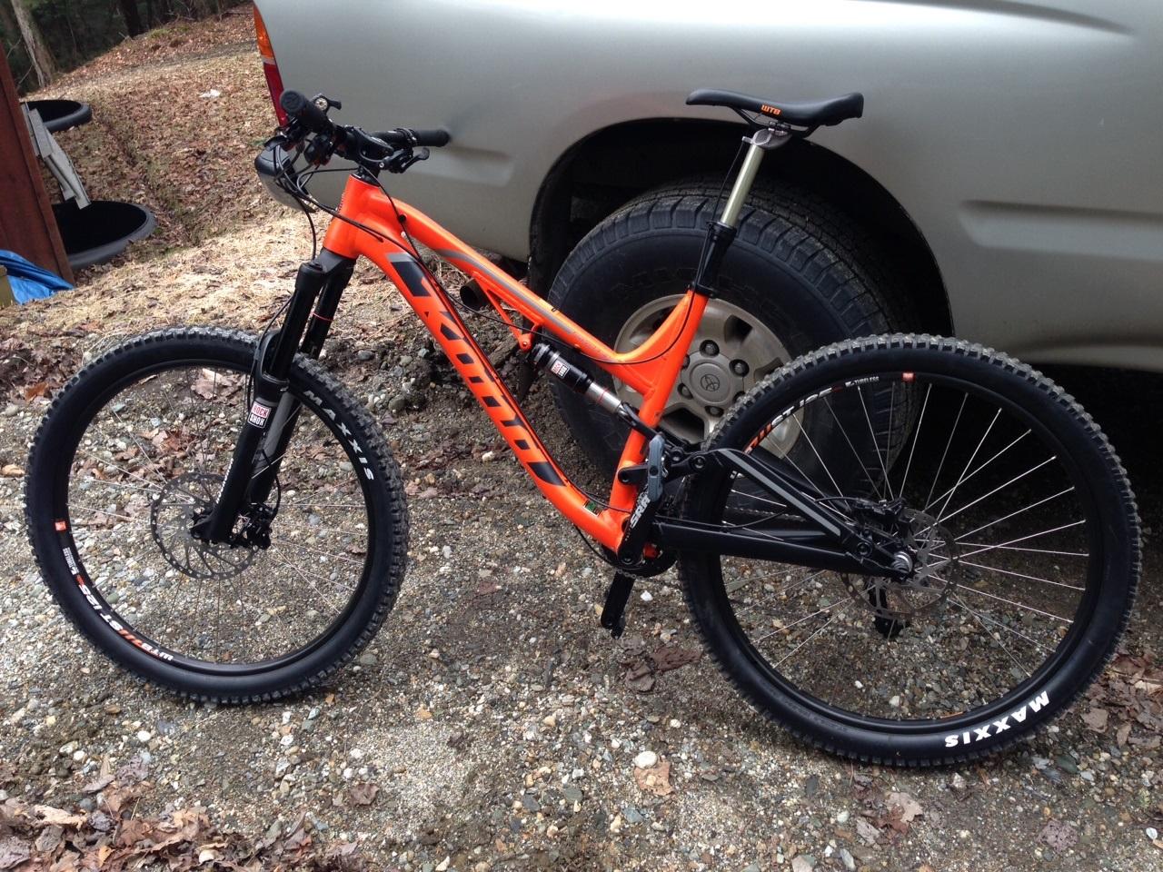 Kona Process 153: A bright orange mountain bike with black accents, positioned on gravel ground next to a silver vehicle. The bike features thick tires with "Maxxis" branding, front suspension forks, and a distinctive aerodynamic frame design. In the background, there are some natural elements and debris, indicating an outdoor setting.