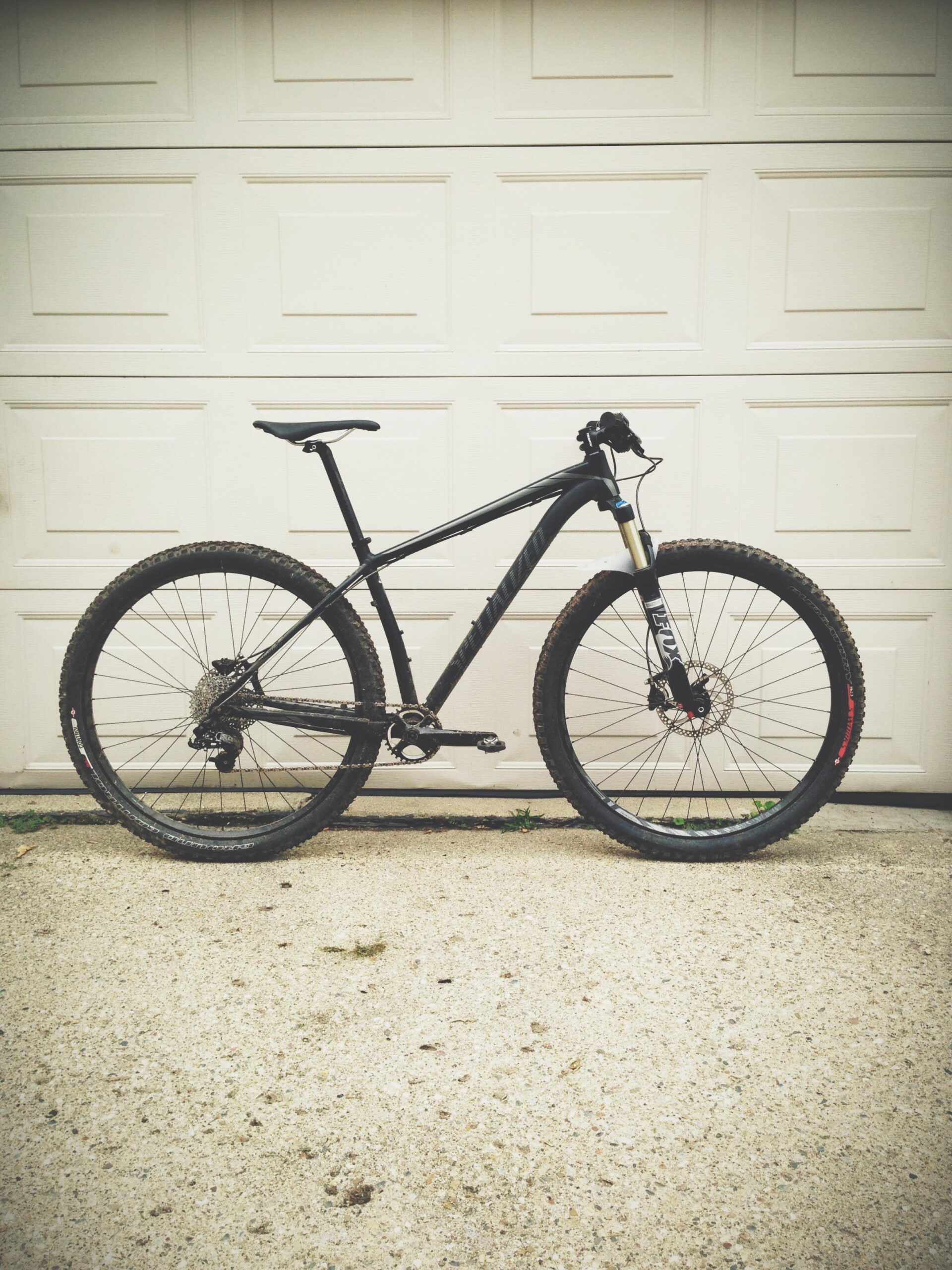 Specialized STUMPJUMPER EVO HT: A black mountain bike with thick tires and a sleek frame, positioned against a white garage door. The bike features a front suspension fork and a geared drivetrain, with some dirt visible on the tires, suggesting recent use.