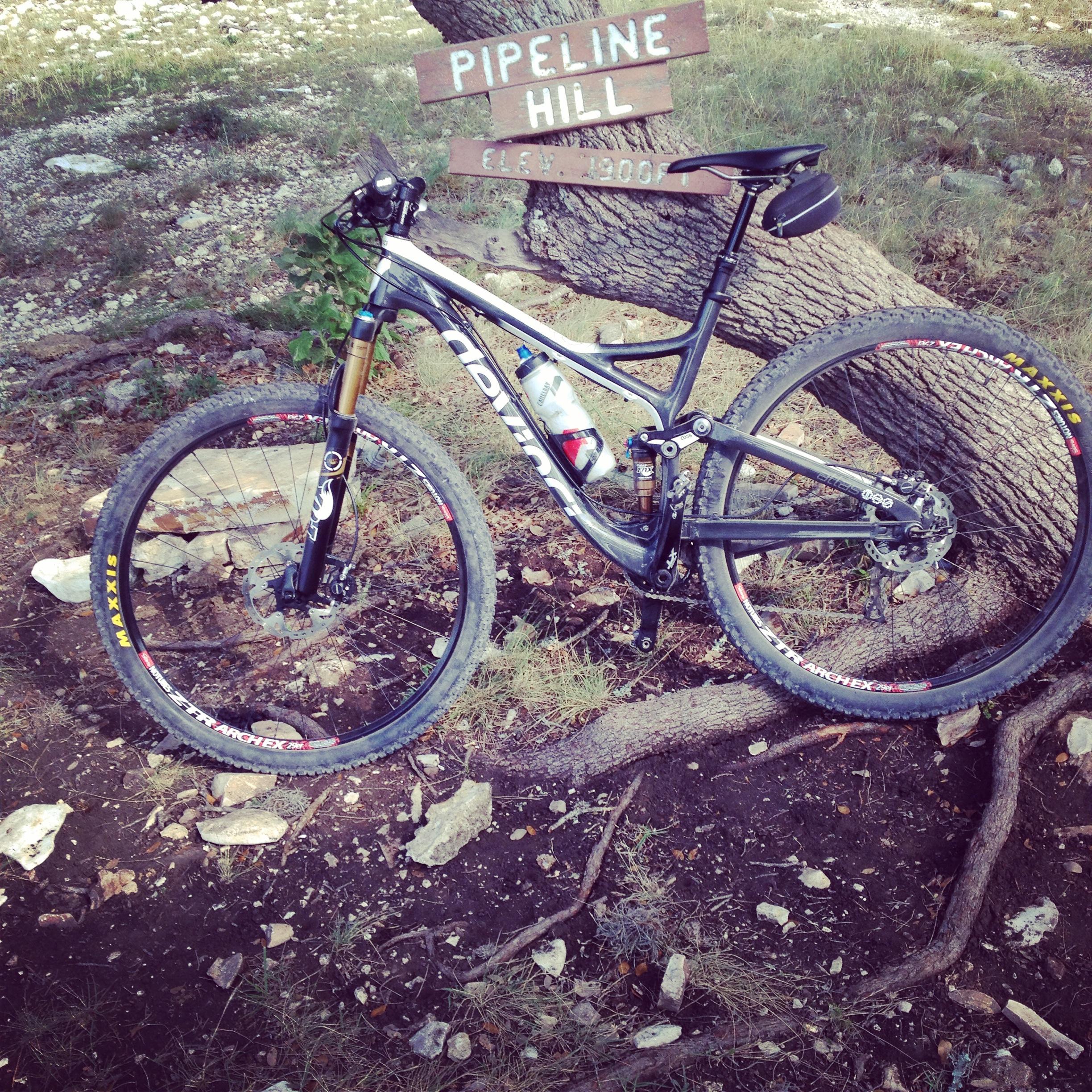 Devinci Atlas Carbon SL: A mountain bike leaning against a tree, with a sign in the background that reads "Pipeline Hill" and indicates an elevation of 1,900 feet. The setting features rocky ground and sparse vegetation, typical of a trail area.