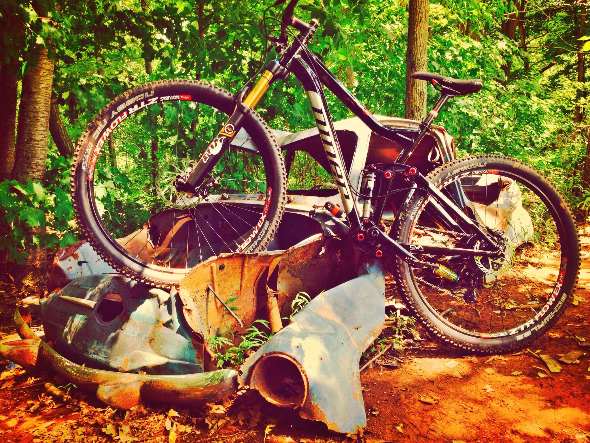 Niner RIP 9: A mountain bike balanced on top of rusted car parts, set against a backdrop of lush green trees and foliage. The bike features thick tires and a sturdy frame, showcasing its rugged design in a natural, overgrown setting.