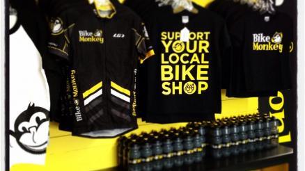 Merchandise display featuring cycling jerseys and t-shirts on hangers, with a prominent black t-shirt that reads, "SUPPORT YOUR LOCAL BIKE SHOP." The backdrop is yellow, and there are bottles arranged neatly at the bottom, all associated with the brand "Bike Monkey."