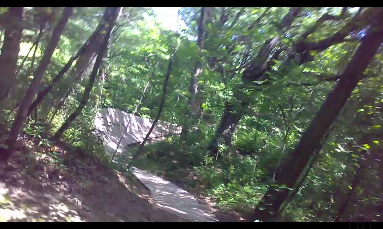 A winding dirt trail through a lush green forest, surrounded by trees and foliage, with a small dirt ramp visible in the distance. The sunlight filters through the leaves, creating a vibrant, natural atmosphere. Outback Trail at Imagination Glenn mountain bike trail.