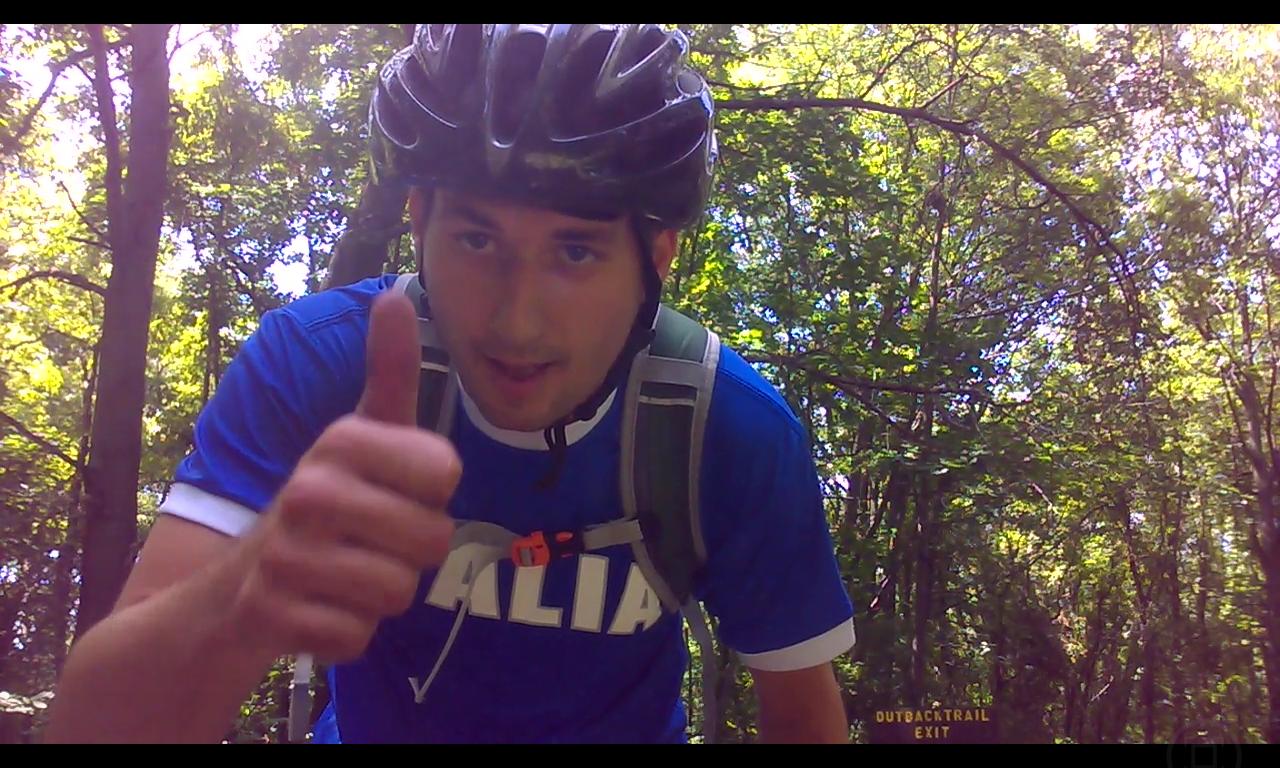 A young man wearing a blue shirt and a helmet gives a thumbs-up while biking on a wooded trail, surrounded by lush greenery. He has a backpack on and is looking directly at the camera, conveying a sense of enthusiasm and adventure. Outback Trail at Imagination Glenn mountain bike trail.