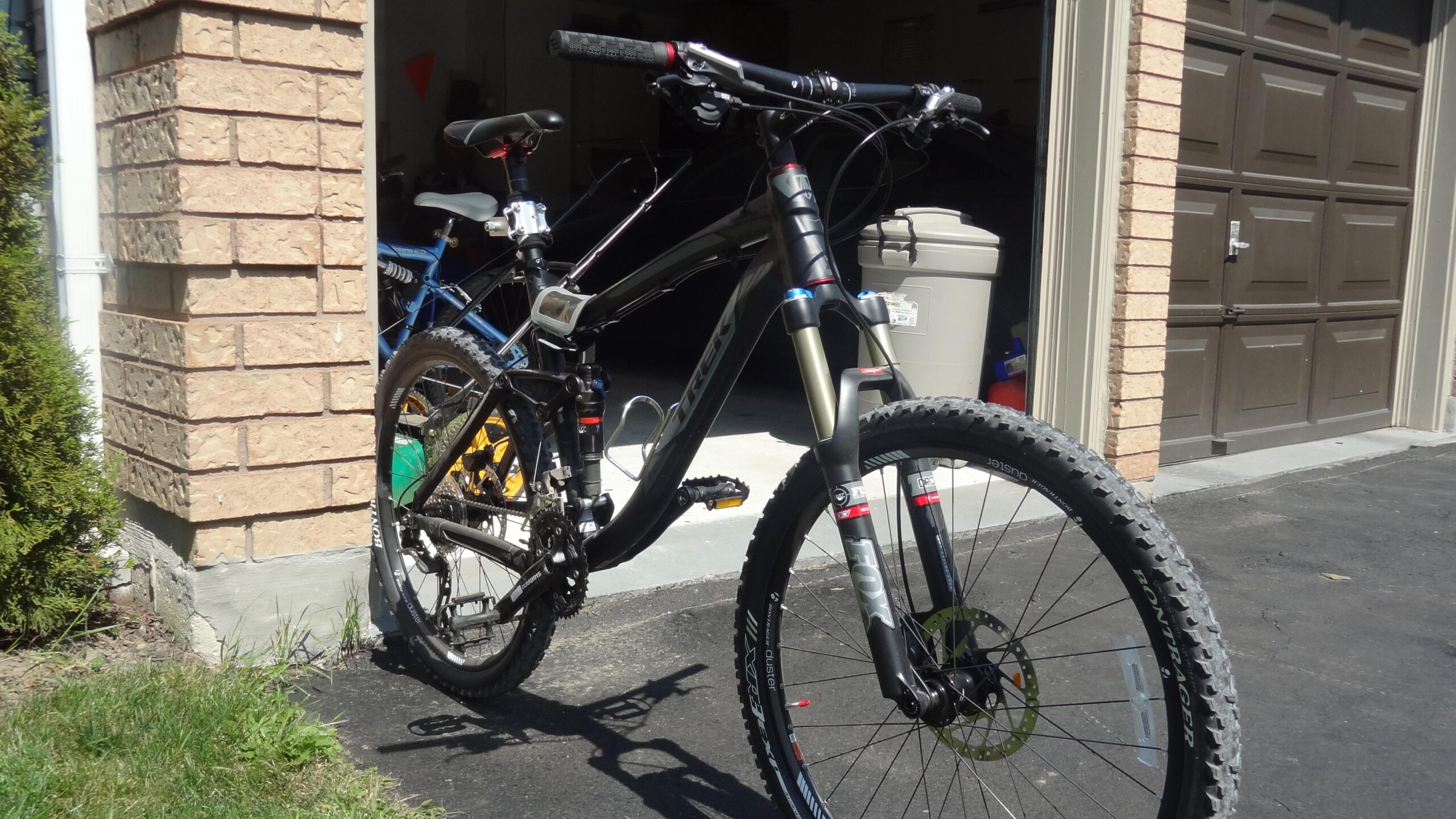 Trek Fuel EX 8: A black mountain bike with front suspension is parked on a driveway next to a house. A blue bike is partially visible in the background, and a garage door is slightly open, revealing items inside. Lush green grass and a bush are present at the edge of the driveway.