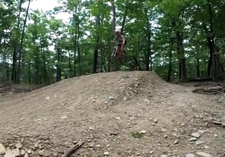 A mountain biker wearing a helmet and riding gear is airborne over a dirt jump in a wooded area, surrounded by green trees. The ground is made up of dirt and small rocks, indicating a biking trail. Carvin's Cove Trail system mountain bike trail.
