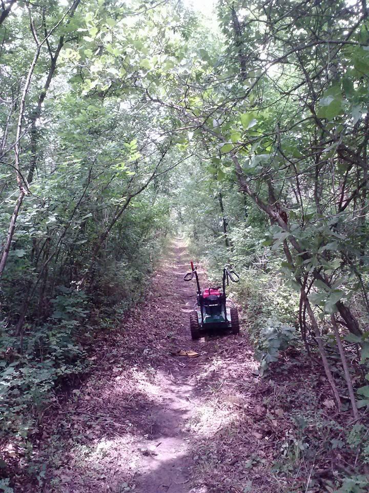 A narrow, dirt path winding through a lush forest with dense green foliage on both sides. In the foreground, a small garden cart is parked on the trail, with a few scattered leaves around it. The scene is bright and natural, conveying a sense of tranquility and exploration. Eagle View, Kaw Lake mountain bike trail.