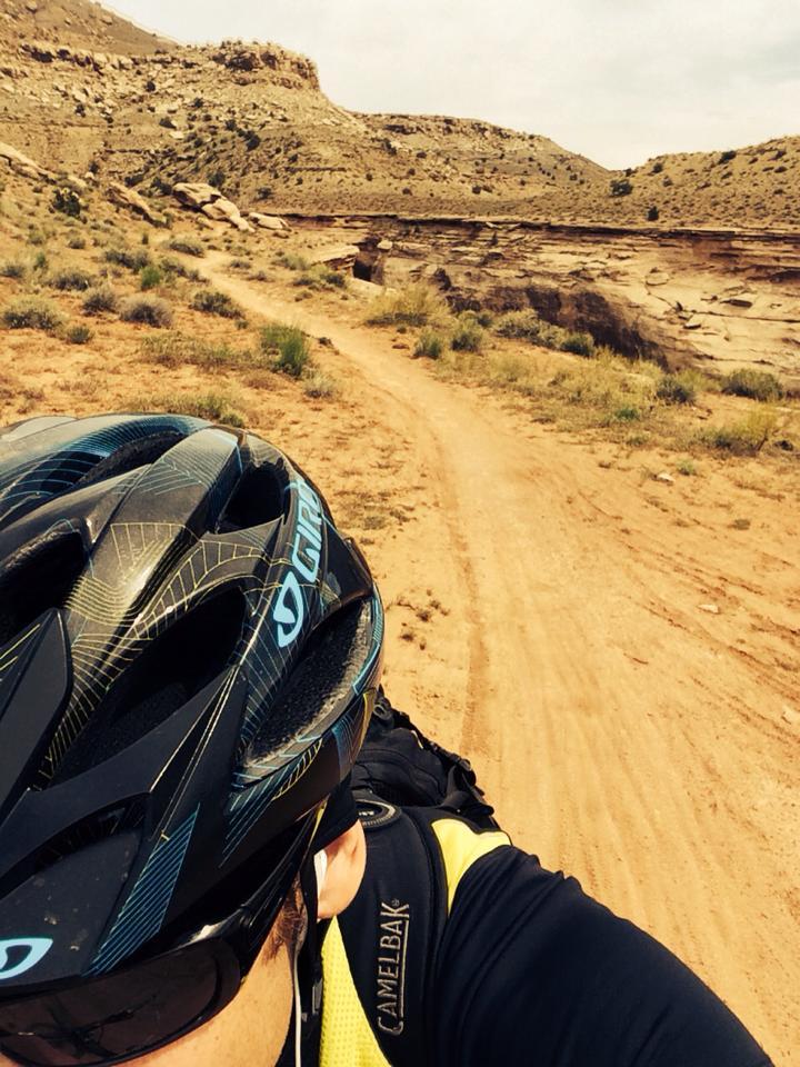 A close-up view of a person wearing a bicycle helmet, with a dirt trail and rocky landscape in the background. The rider is dressed in a cycling jersey, and a CamelBak hydration pack is visible on their back. The scene is set in a rugged outdoor environment, suggesting mountain biking or trail riding. Kokopelli Trail mountain bike trail.