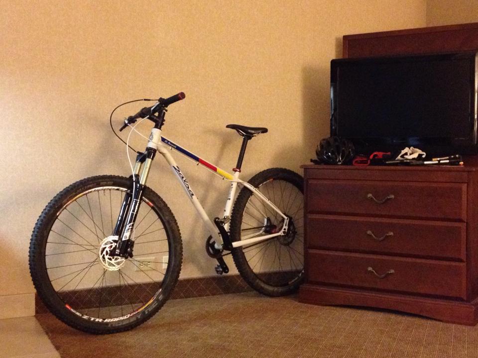 A mountain bike leaning against a dresser in a hotel room, with a television on top of the dresser and cycling gear, including a helmet and gloves, placed nearby. The room features neutral-colored walls and carpet.