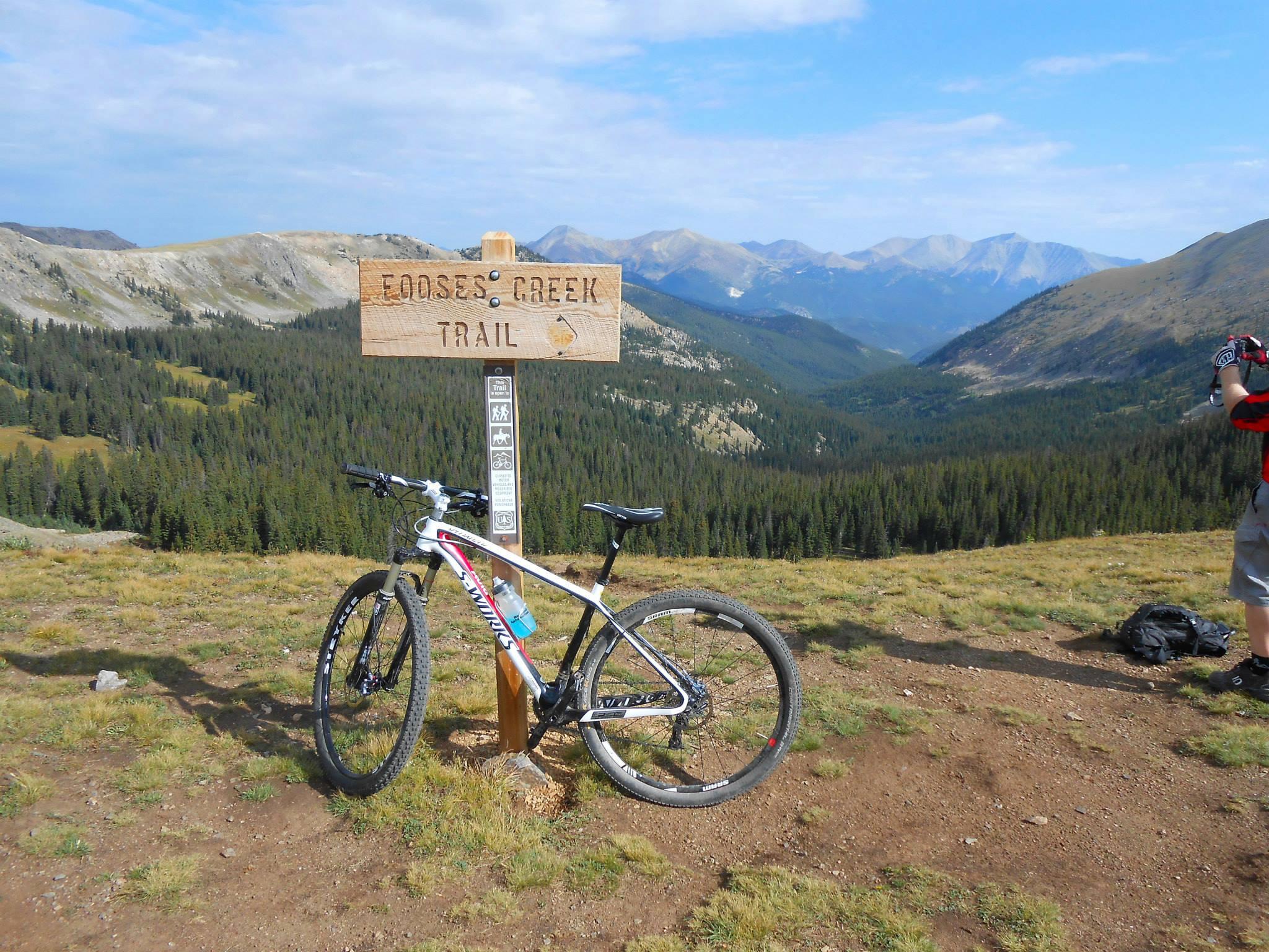 Specialized Stumpjumper Comp Carbon 29: A mountain bike parked next to a wooden sign indicating the start of the Eootses Creek Trail, with a scenic view of mountainous terrain and forests in the background. A person is visible in the distance, capturing the landscape. The sky is partly cloudy, and the environment showcases rich greenery.