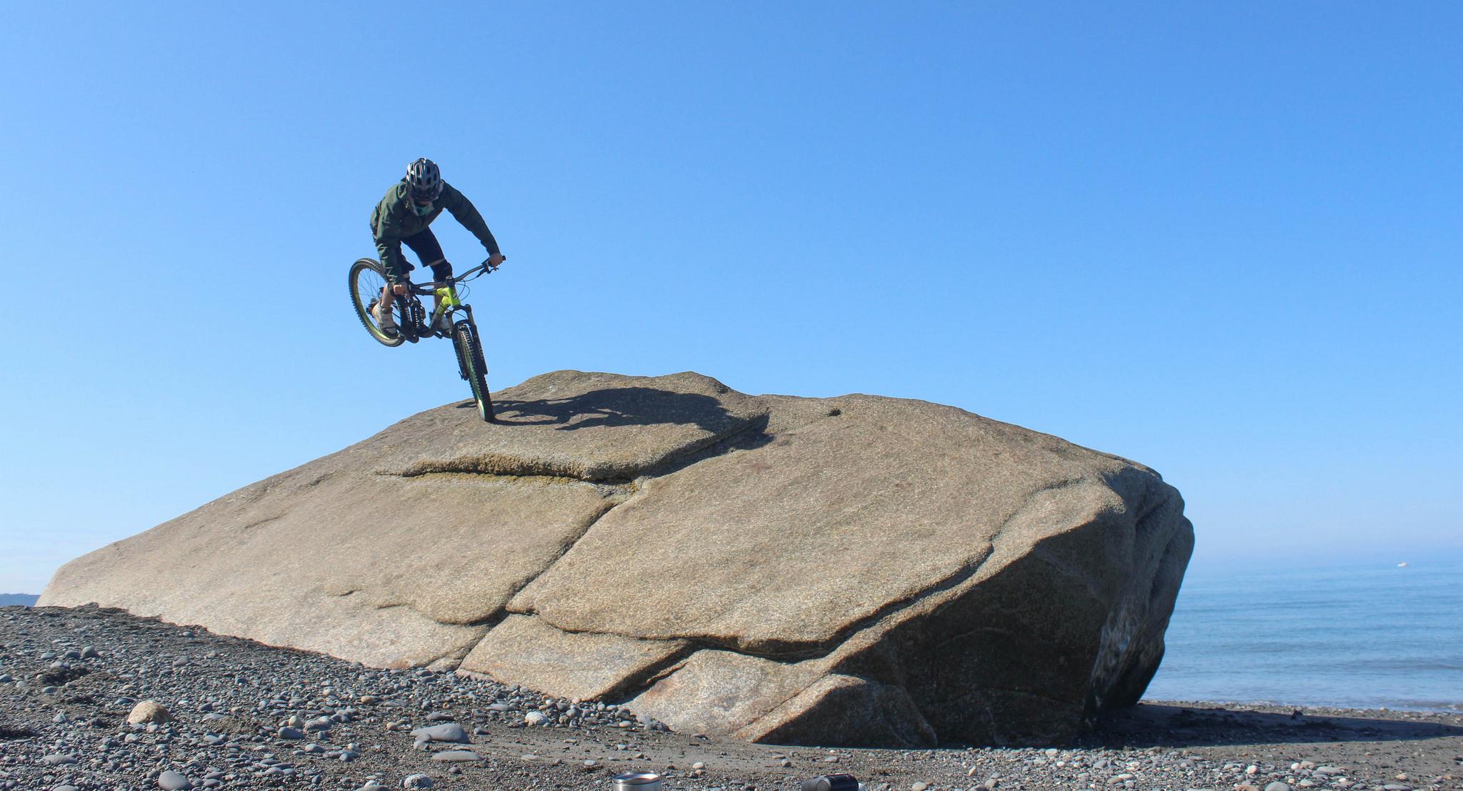 A mountain biker performing a stunt on a large rock by the beach, with a clear blue sky and ocean in the background. The biker is wearing a helmet and has one wheel lifted off the rock, showcasing a dynamic and adventurous moment. Deep creek beach mountain bike trail.