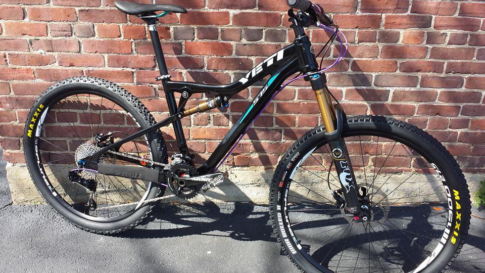 Yeti 575: A black mountain bike with colorful accents stands against a brick wall. The bike features thick tires with Maxxis branding, a dual suspension system, and a sleek frame design, showcasing its suitability for off-road biking. The sunlight highlights the bike's components, emphasizing the gold fork and colorful details.