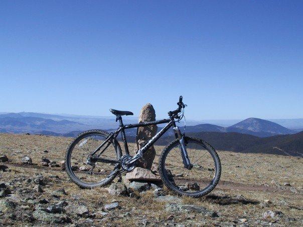 A black mountain bike leaning against a large stone in a rocky, mountainous landscape under a clear blue sky. The background features distant hills and valleys, showcasing a scenic outdoor environment. Monarch Crest Trail mountain bike trail.
