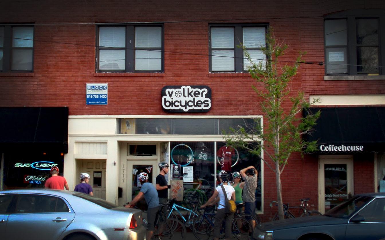 A bicycle shop named "Volker Bicycles" is depicted in this image, featuring a brick exterior with large windows. Several cyclists wearing helmets are gathered near the entrance, with bicycles parked outside. To the right of the shop, there is a coffeehouse with a black awning. A car is parked in front of the shop, and a sign for another business can be seen overhead.