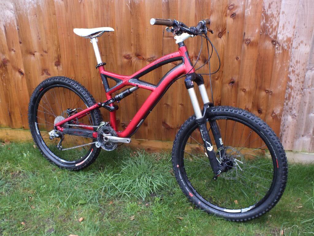 A red mountain bike with a sturdy frame, equipped with wide tires and front suspension, positioned on grass in front of a wooden fence. The bike features a white saddle, black handlebars, and a visible drivetrain.