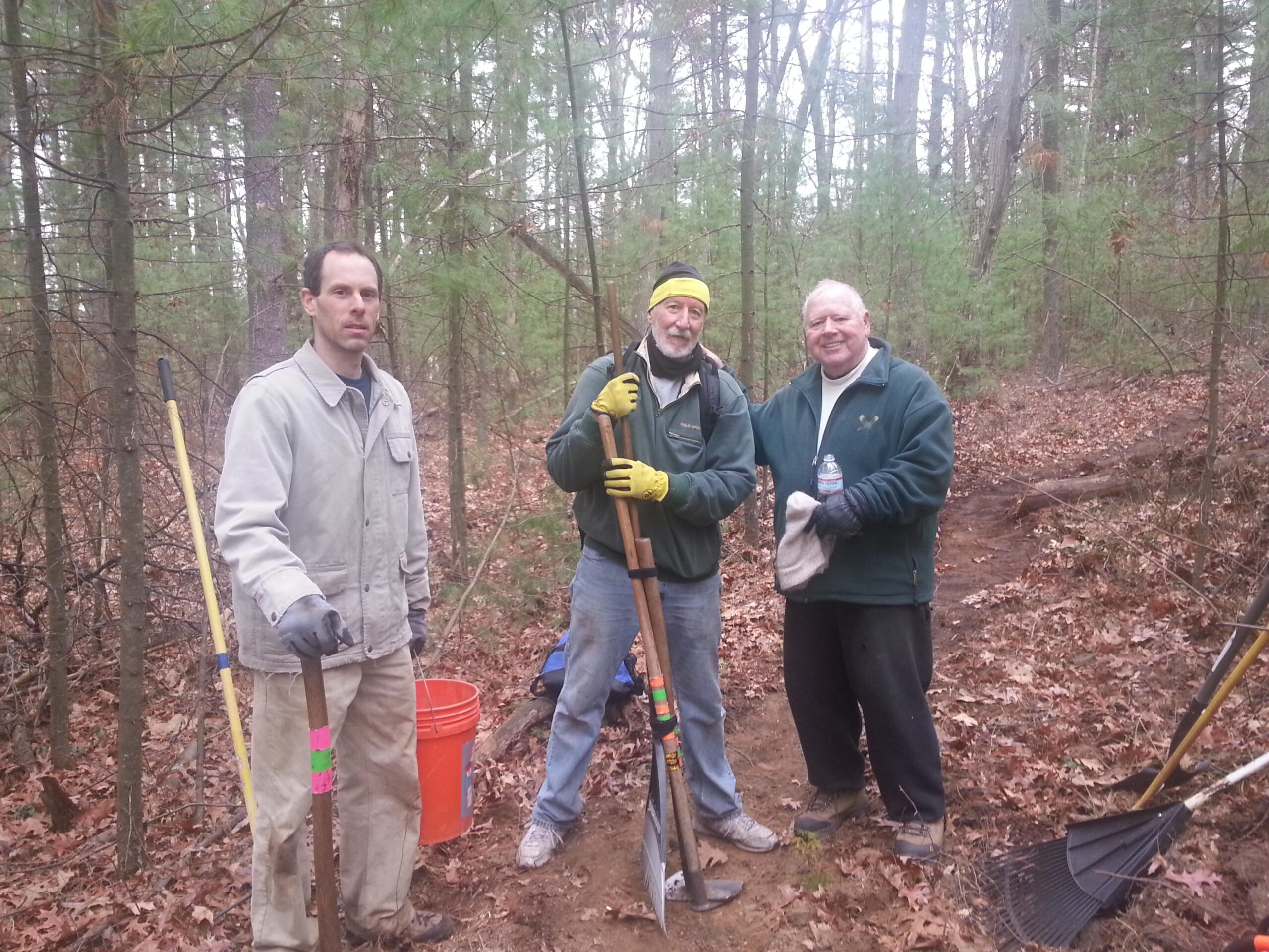 Three men standing in a wooded area, wearing work gloves and casual outdoor clothing. They are holding gardening tools, including shovels and rakes. The background features trees and fallen leaves, indicating a natural setting. One man smiles while another holds a water bottle, suggesting they are engaged in outdoor work or volunteering. A plastic bucket is positioned nearby. Russell Mill mountain bike trail.