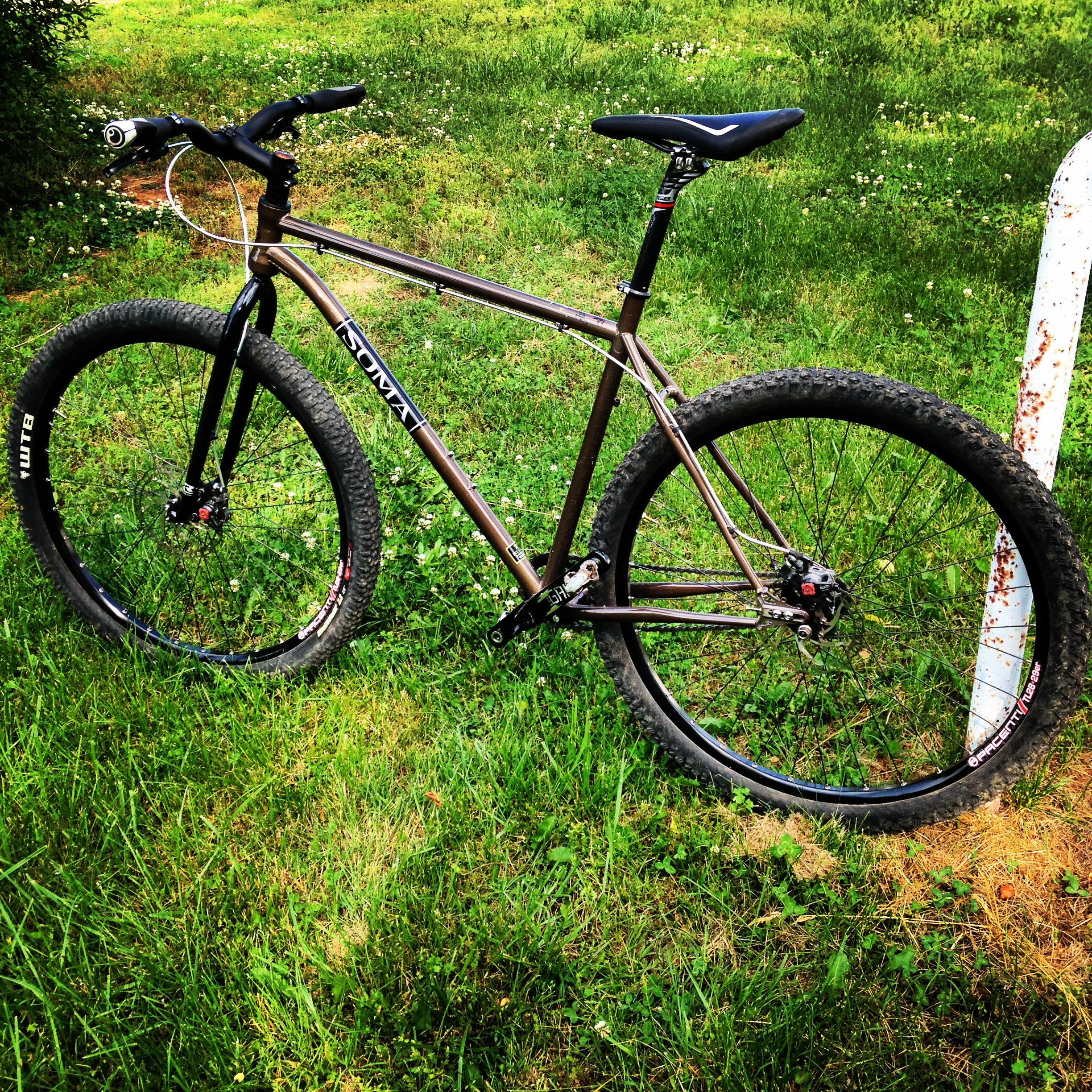 Soma Juice (Brownstone): A brown mountain bike resting on green grass, featuring thick tires and a sleek frame. The bike has a black seat and handlebar grips, with a visible cable attached to the front. Nearby, a rusty white pole can be seen in the background.