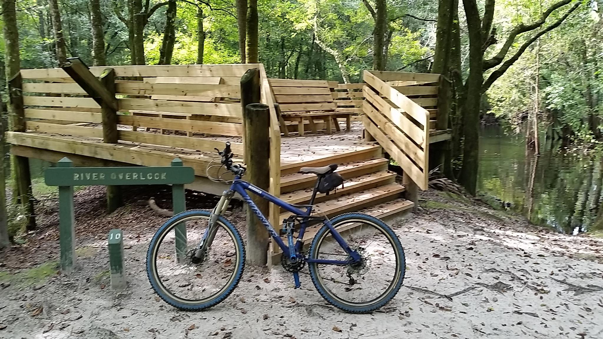 Turner 5.Spot: A blue mountain bike is parked next to a wooden overlook platform by a river, surrounded by lush green trees. A sign reads "River Overlook," and the sandy ground is scattered with leaves, indicating a natural, serene setting.