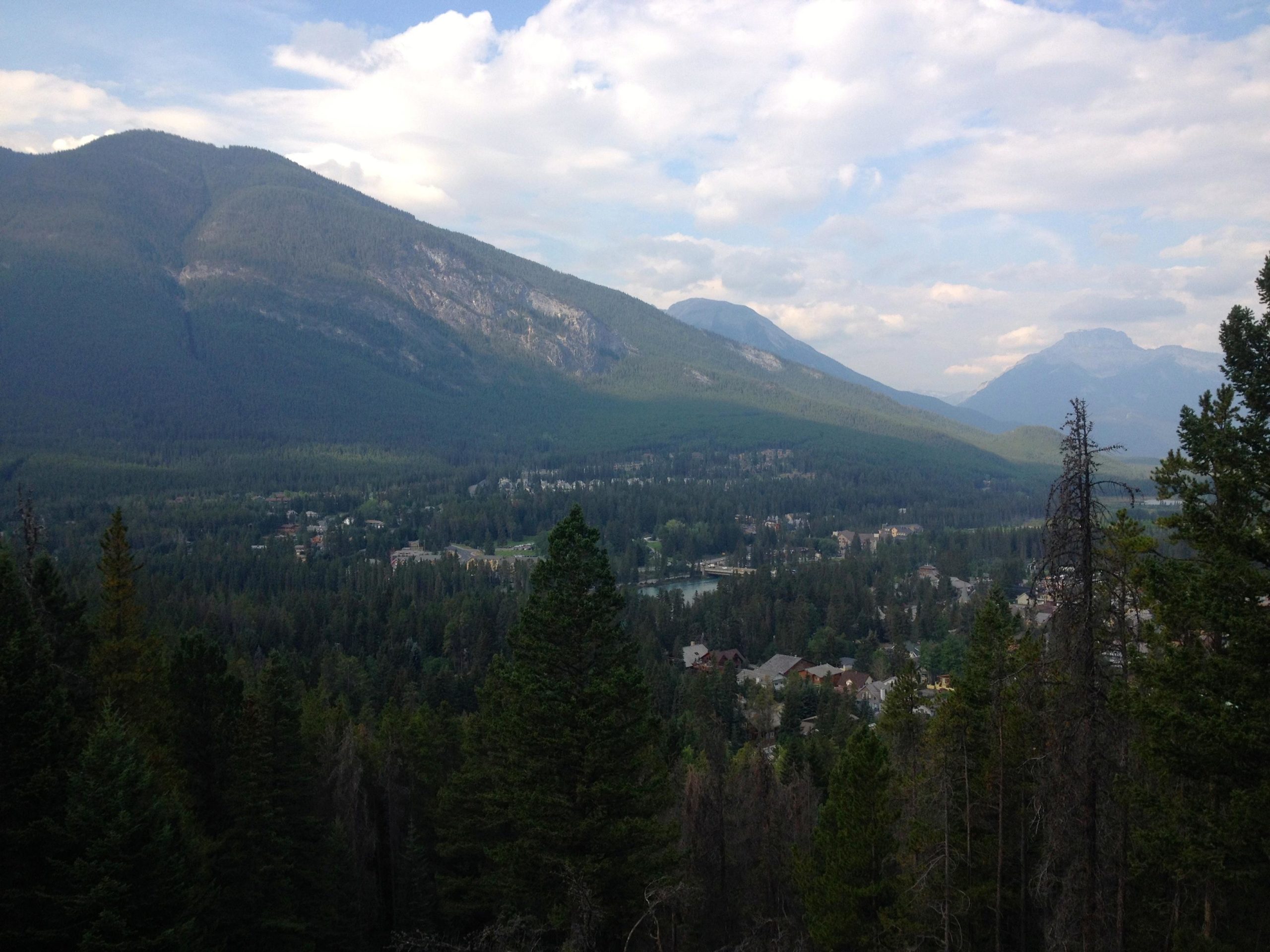 A panoramic view of a mountainous landscape with lush green forests, showcasing distant peaks under a partly cloudy sky. In the foreground, evergreen trees frame the scene, while a small town nestled among the trees can be seen in the valley below. Stoney Squaw Upper & Lower mountain bike trail.