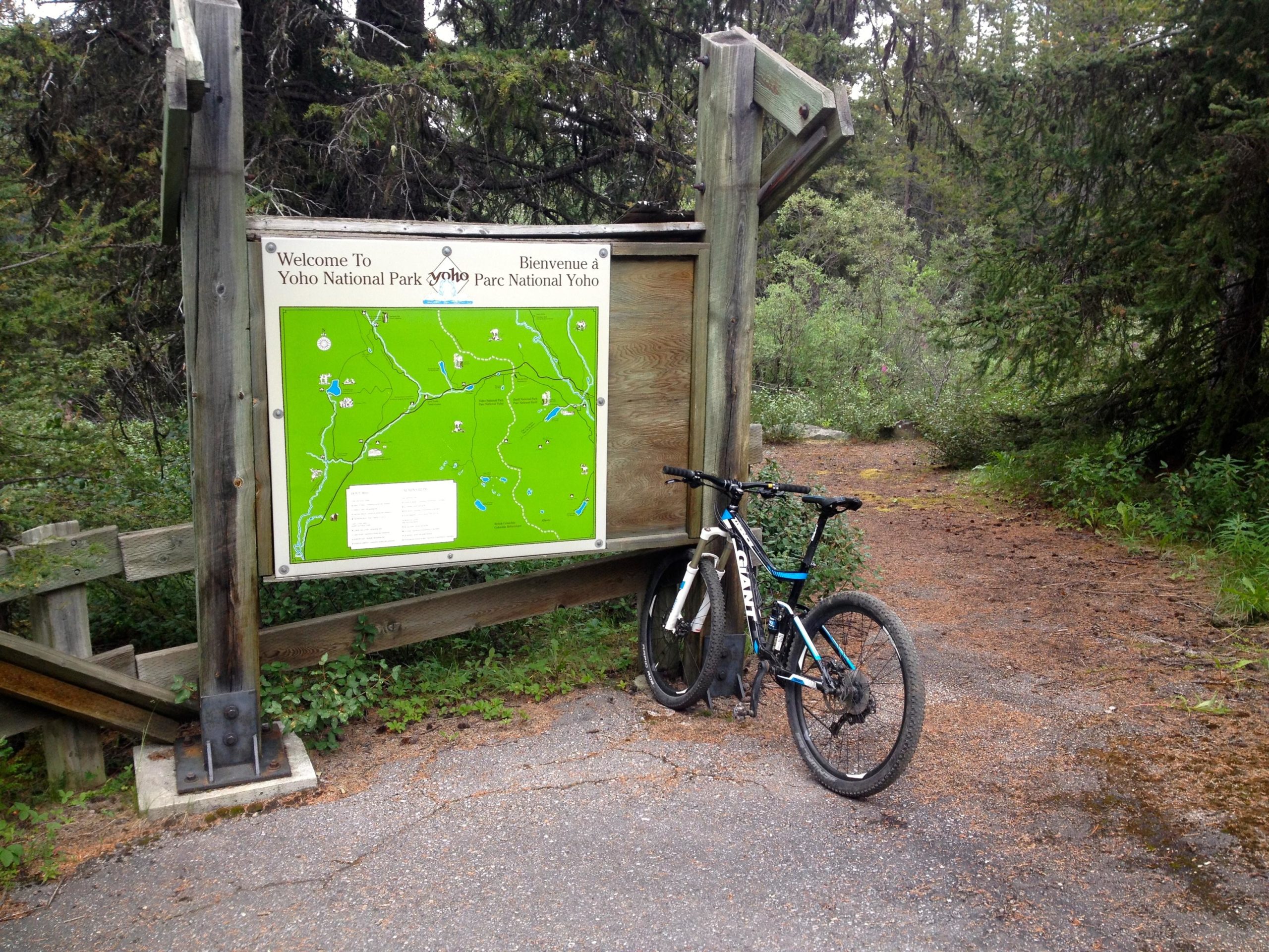 A welcome sign and trail map for Yoho National Park, surrounded by lush greenery, with a mountain bike leaning against the sign. The pathway is visible leading into the forested area. Great Divide Bike Path mountain bike trail.