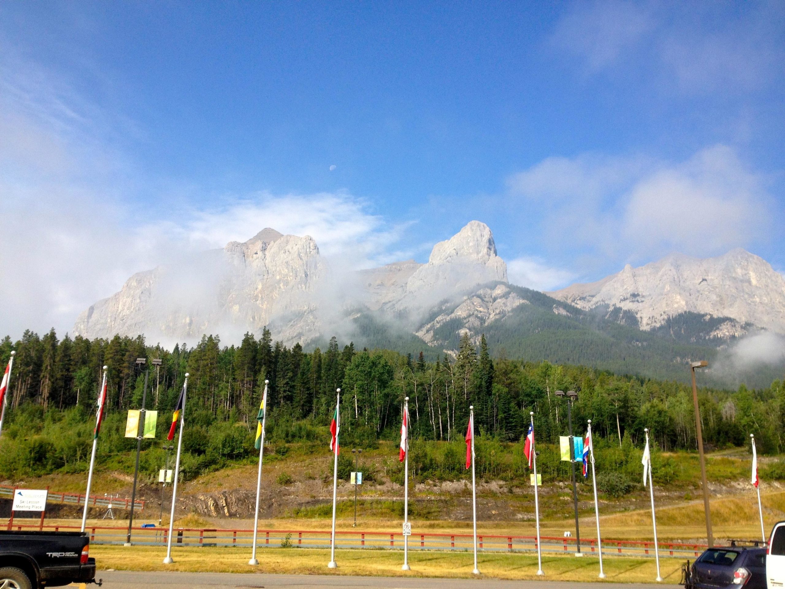 A scenic view of towering mountains partially shrouded in mist, set against a blue sky. In the foreground, a row of flags from various countries is prominently displayed, with a green landscape and trees behind them. The image captures the natural beauty of the mountains and the welcoming atmosphere created by the flags. Canmore Nordic Centre mountain bike trail.