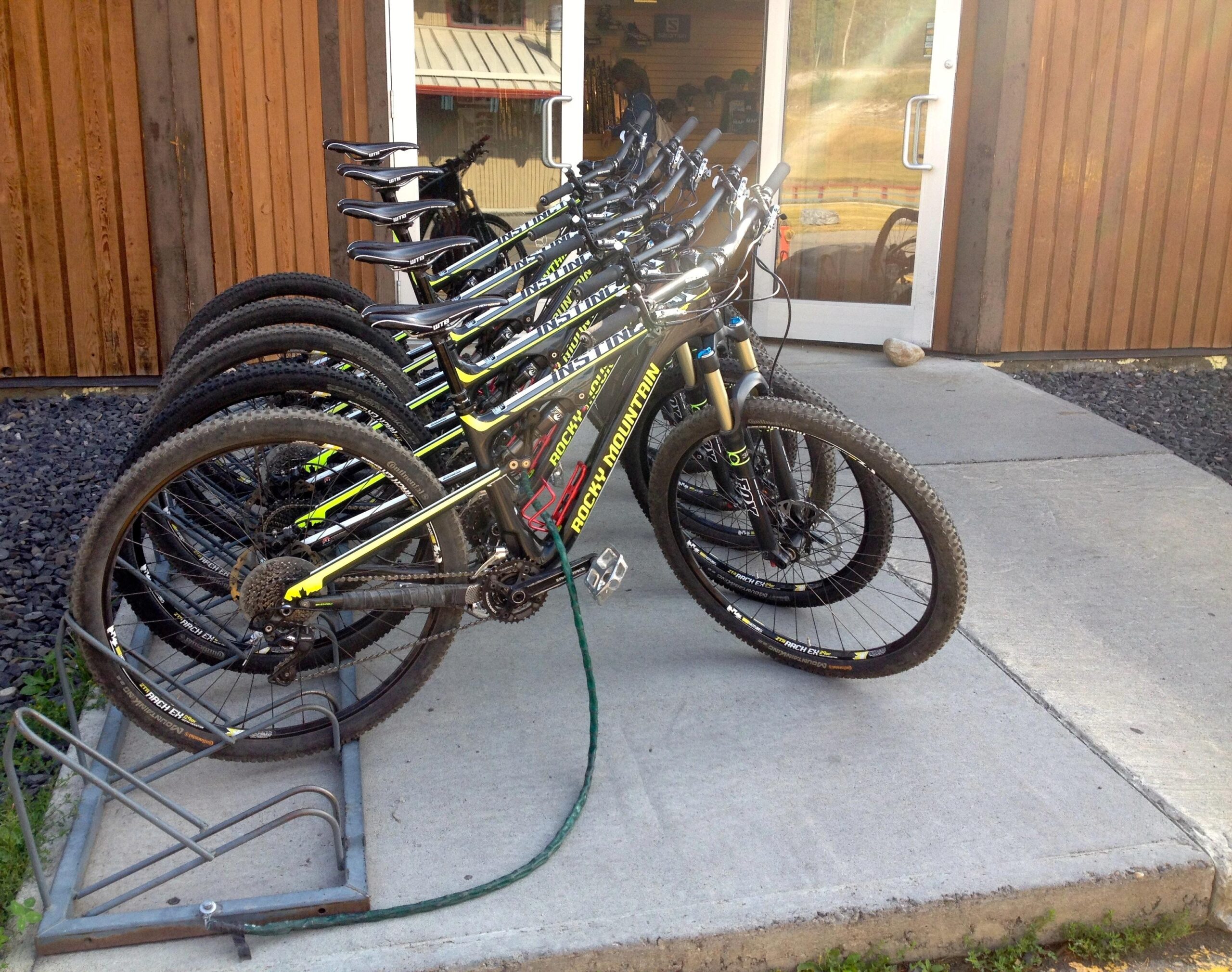 A row of mountain bikes secured in a bike rack in front of a wooden building. The bikes are positioned with their handlebars facing outward, showcasing their distinctive designs. The ground is made of concrete, and the setting appears to be a bike rental or shop area. Canmore Nordic Centre mountain bike trail.
