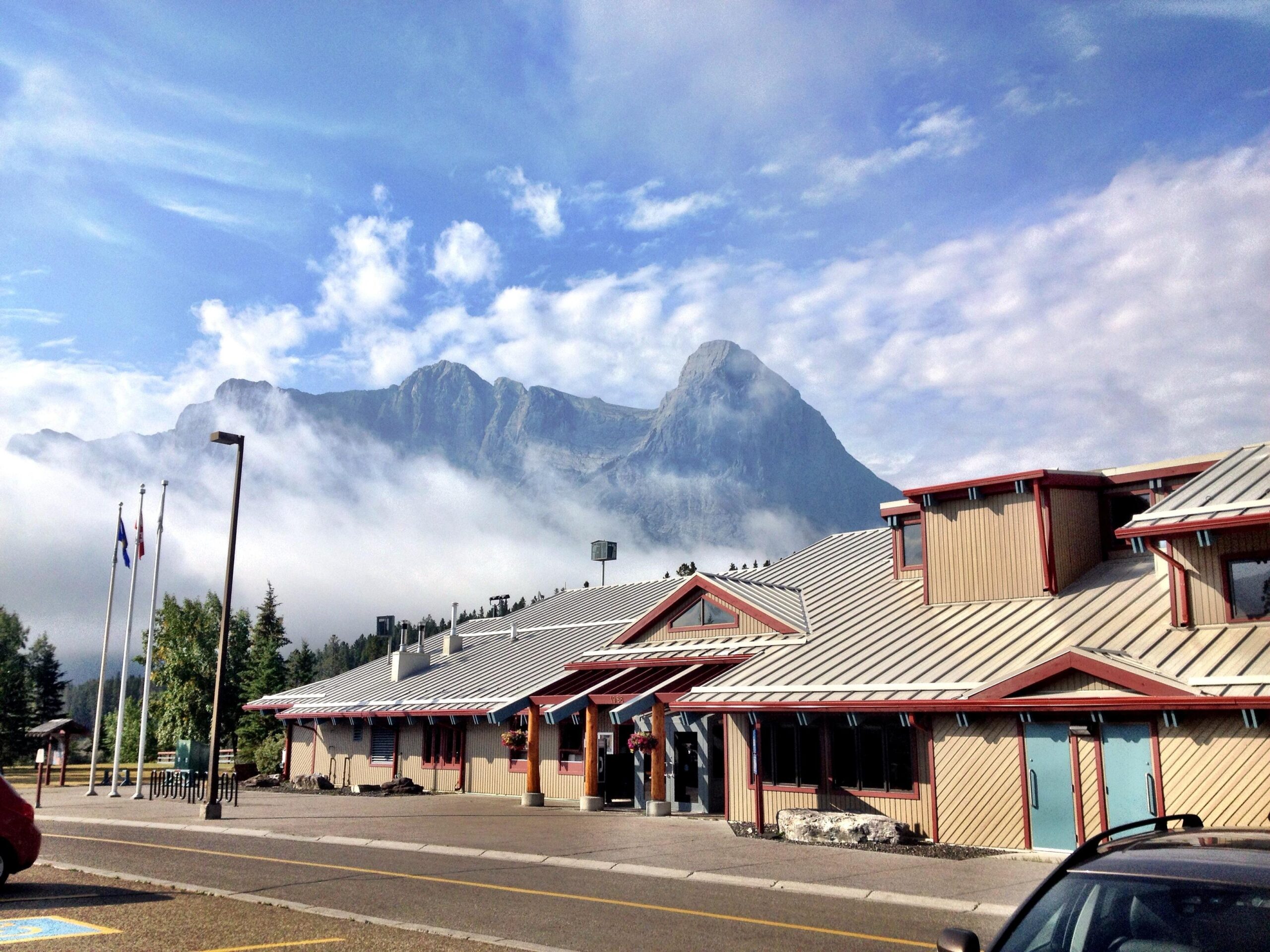 A serene landscape featuring a building with a red-roofed design in the foreground, surrounded by trees and a paved road. Behind the building, majestic mountains rise, partially shrouded in clouds under a blue sky with scattered clouds. Two flagpoles stand in front of the building, adding to the tranquil scene. Canmore Nordic Centre mountain bike trail.