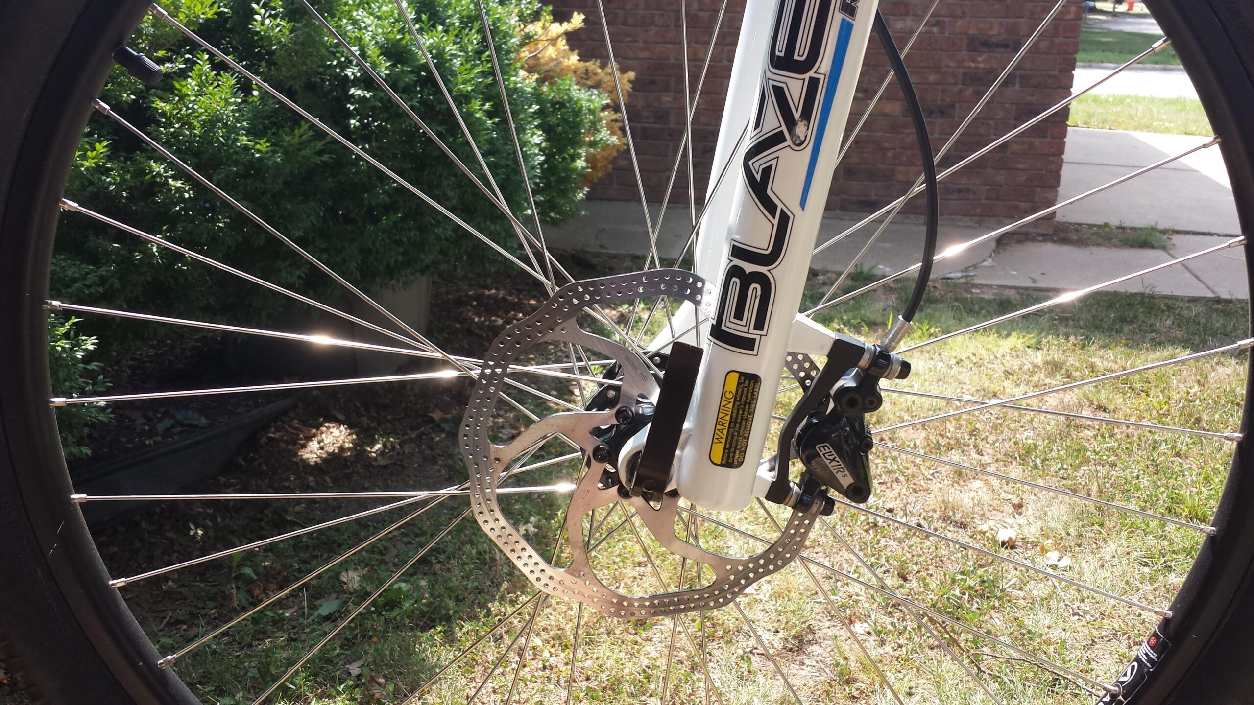 Specialized Hardrock Disc 29: Close-up view of a bicycle wheel featuring a white frame with the word "BLAZE" visible, disc brake system, and metal spokes, set against a background of green shrubs and grass.