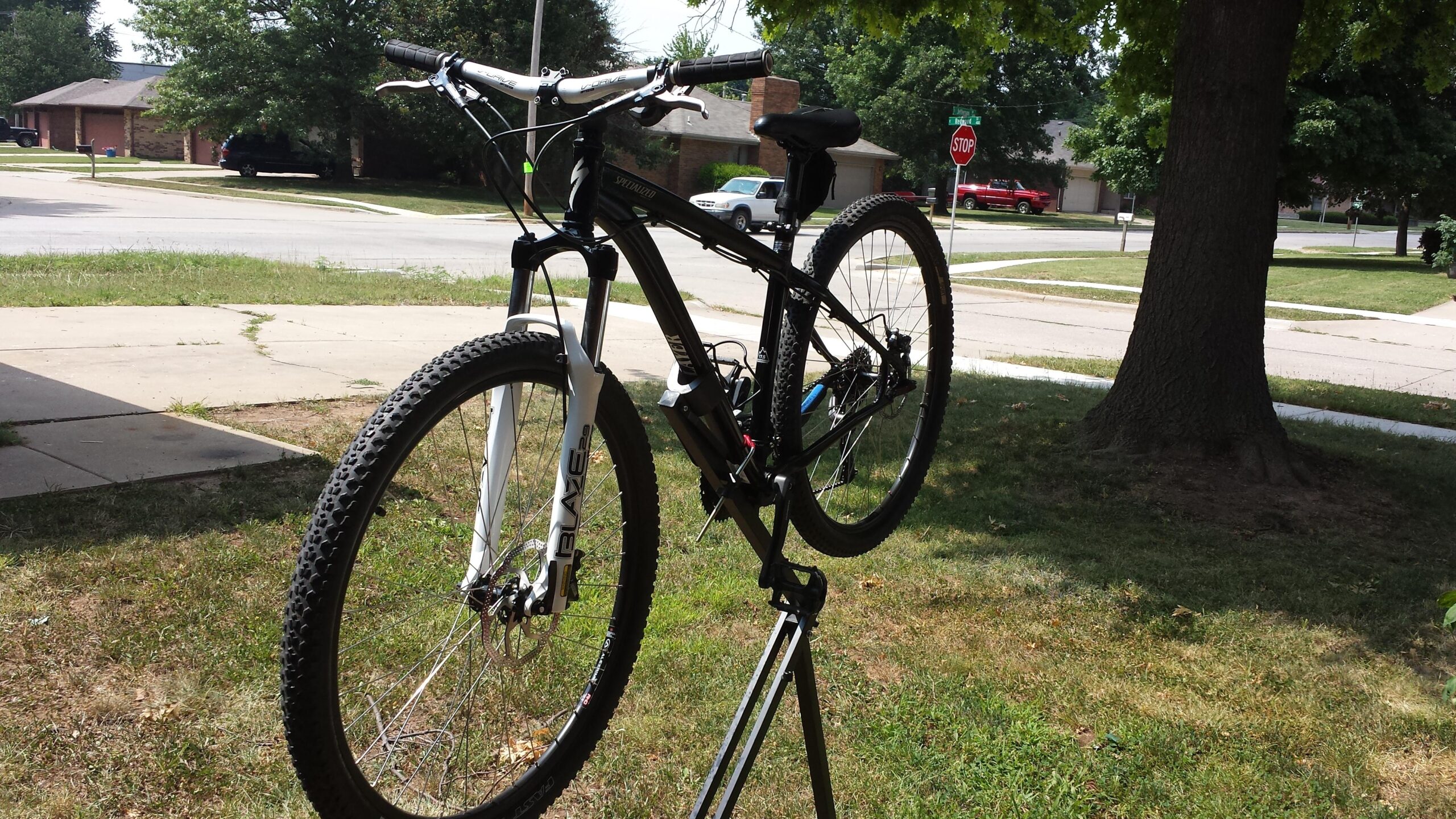 Specialized Hardrock Disc 29: A black and white mountain bike is propped on a maintenance stand in a grassy front yard. The background features residential houses, a stop sign, and trees.