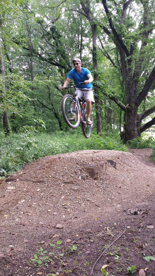 A person in a blue shirt and shorts is performing a jump on a mountain bike over a dirt ramp, surrounded by lush greenery and trees. The cyclist is airborne with both wheels off the ground, showcasing a dynamic biking action. Bourissa Hills mountain bike trail.