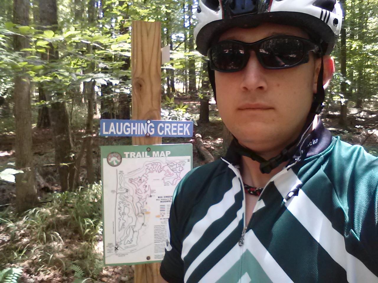 A person wearing a cycling jersey and sunglasses stands near a sign for Laughing Creek, which includes a trail map, surrounded by a wooded area with trees and foliage. Big Creek mountain bike trail.