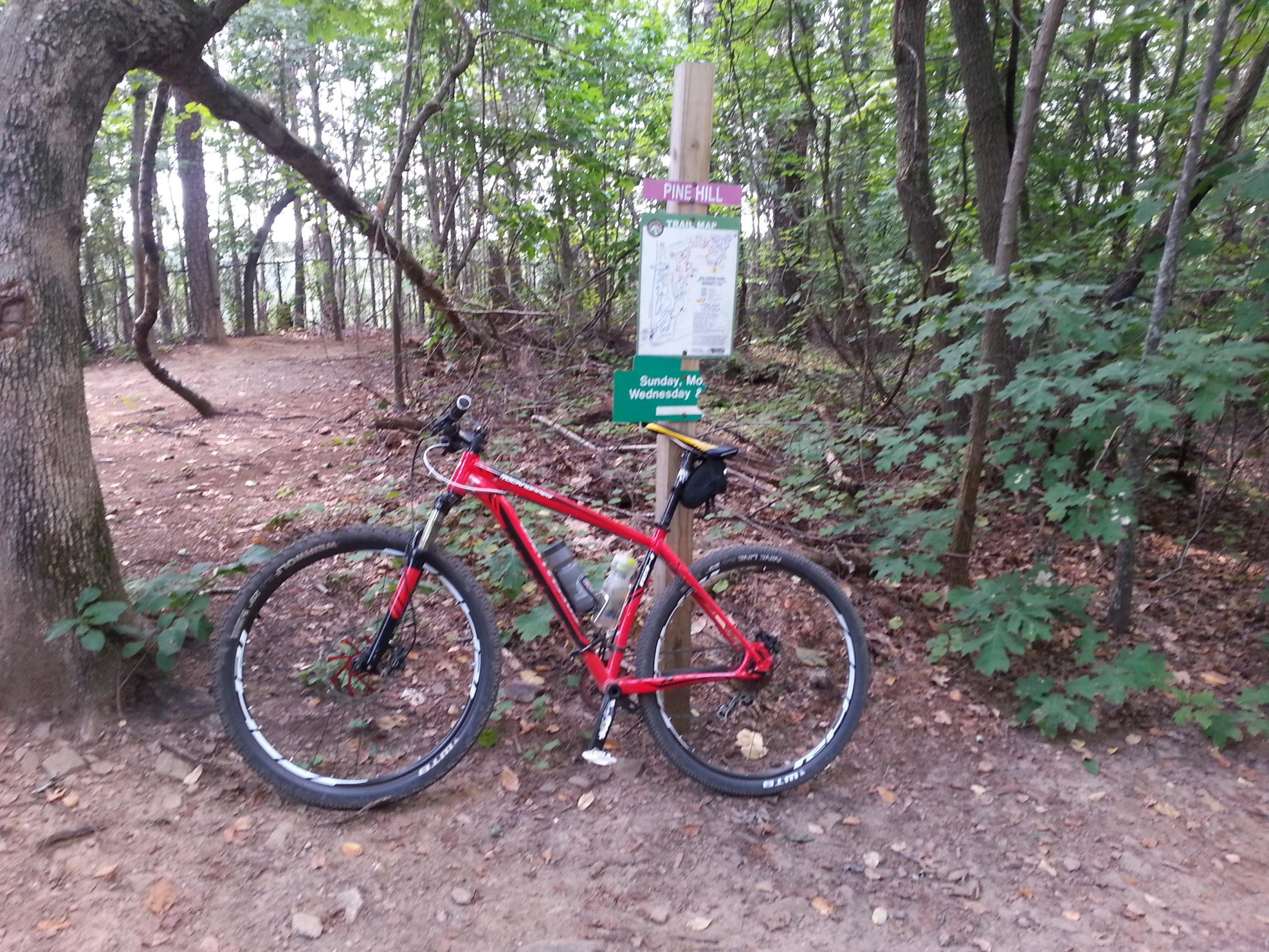 A red mountain bike leaning against a wooden trail sign in a wooded area. The sign displays a map of the Pine Hill trail. Surrounding the bike are trees and shrubs, with a dirt path leading into the forest. Big Creek mountain bike trail.
