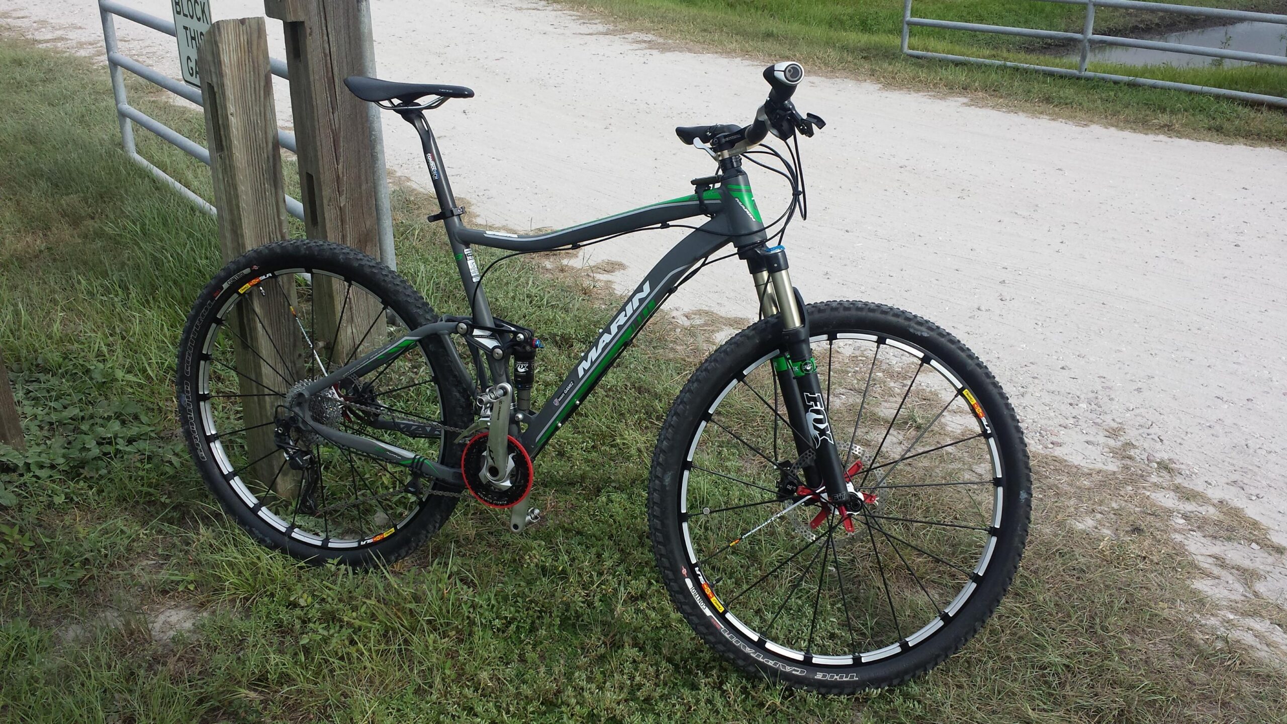 Marin Rift Zone 29er XC8: Mountain bike leaning against a wooden post near a gravel path, surrounded by grass. The bike features a gray frame with green accents and black components, showcasing thick tires designed for off-road terrain. A sign in the background warns not to block the gate.
