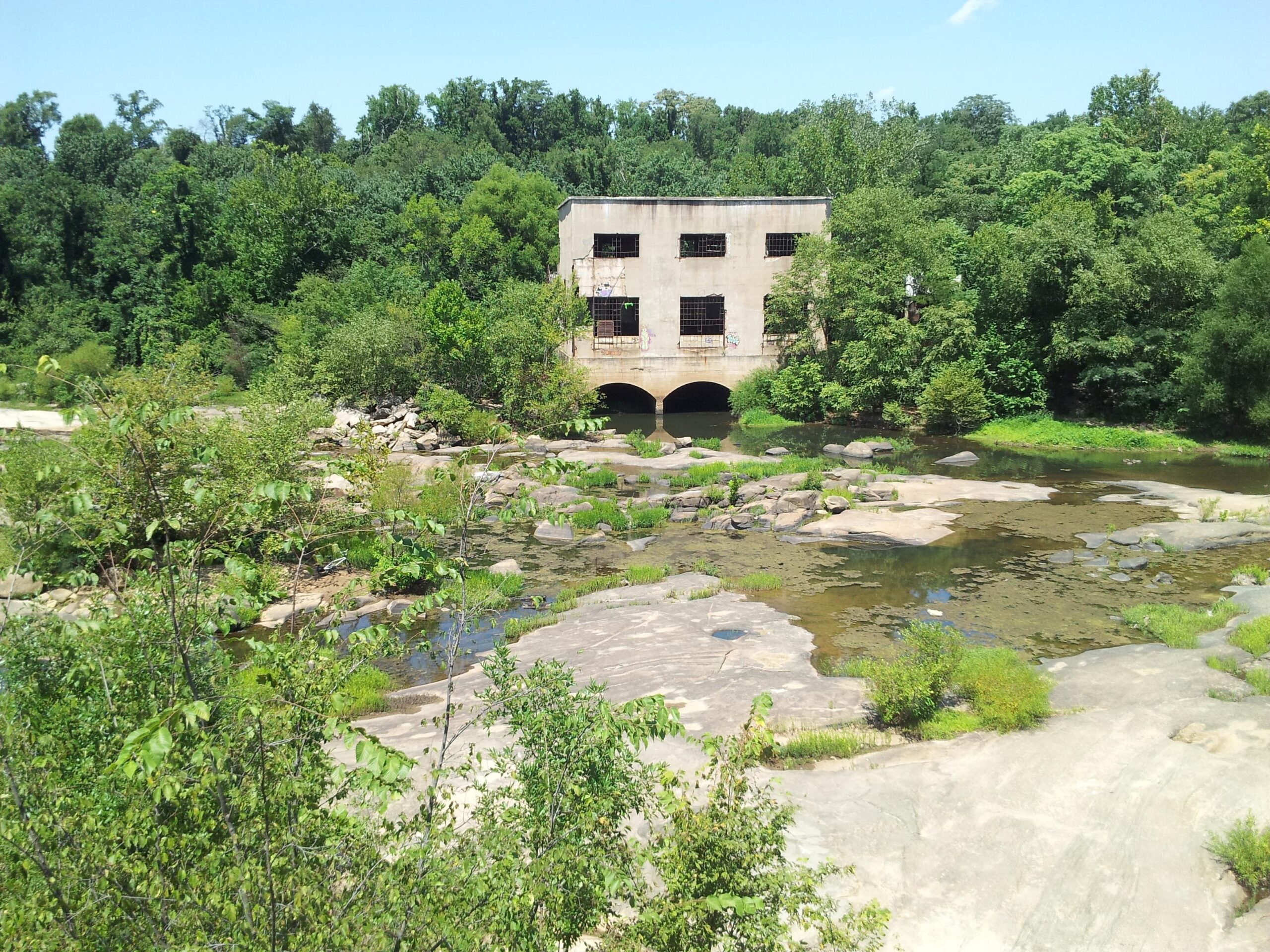 Abandoned concrete building surrounded by lush greenery and rocky terrain, with a shallow stream reflecting the blue sky. Belle Isle mountain bike trail.