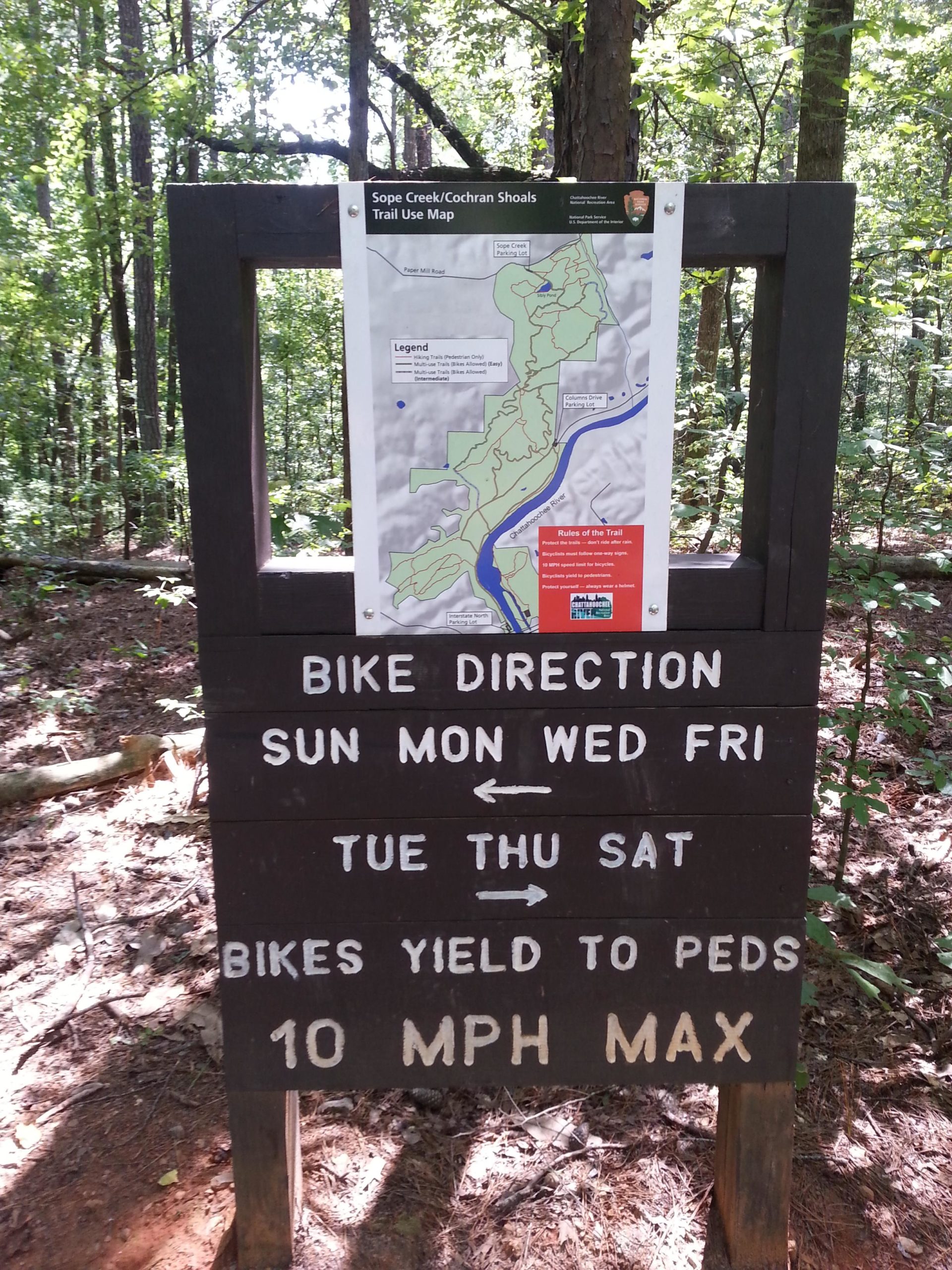 A wooden signpost in a wooded area features a trail use map for Sope Creek/Cochran Shoals. The sign includes directions for biking, specifying designated biking days of the week, a note that bikes yield to pedestrians, and a maximum speed limit of 10 MPH. Sope Creek mountain bike trail.