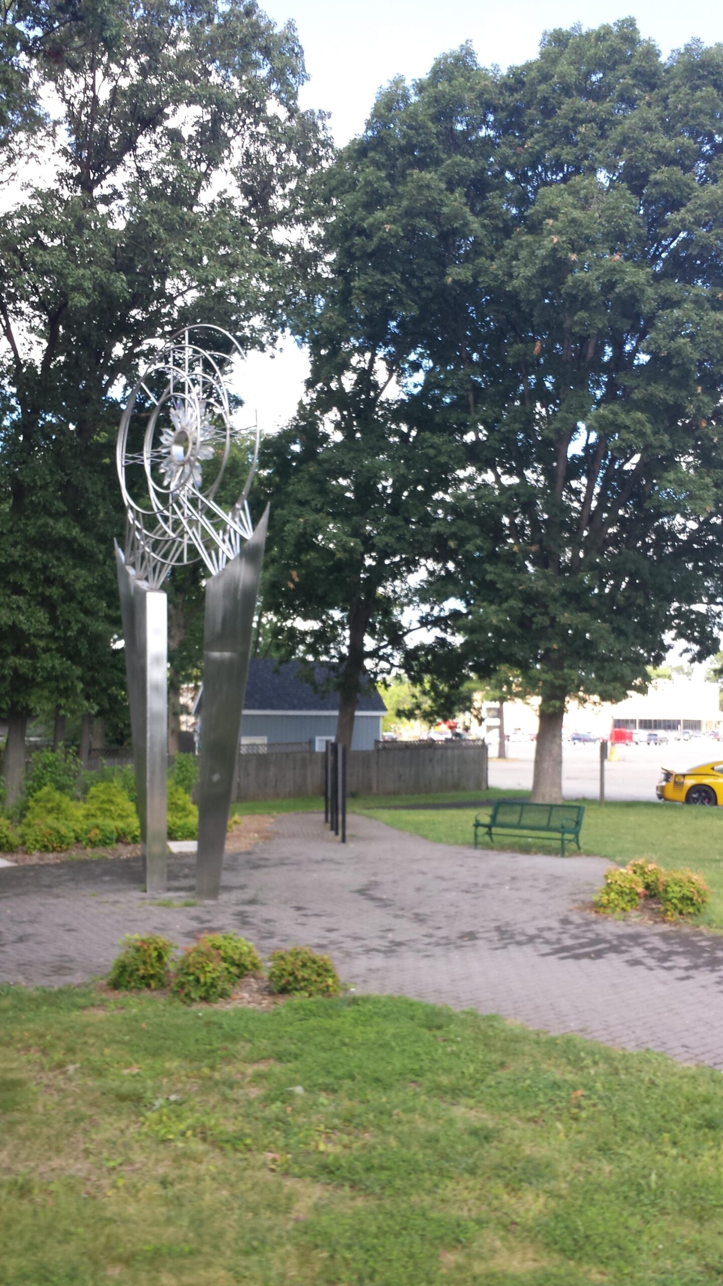 Trek 4500: A modern metallic sculpture stands in a park, surrounded by green grass and shrubs. In the background, tall trees provide shade, and a park bench is positioned nearby. A yellow car is visible in the background, along with a glimpse of a building beyond the trees. The scene appears peaceful and inviting.