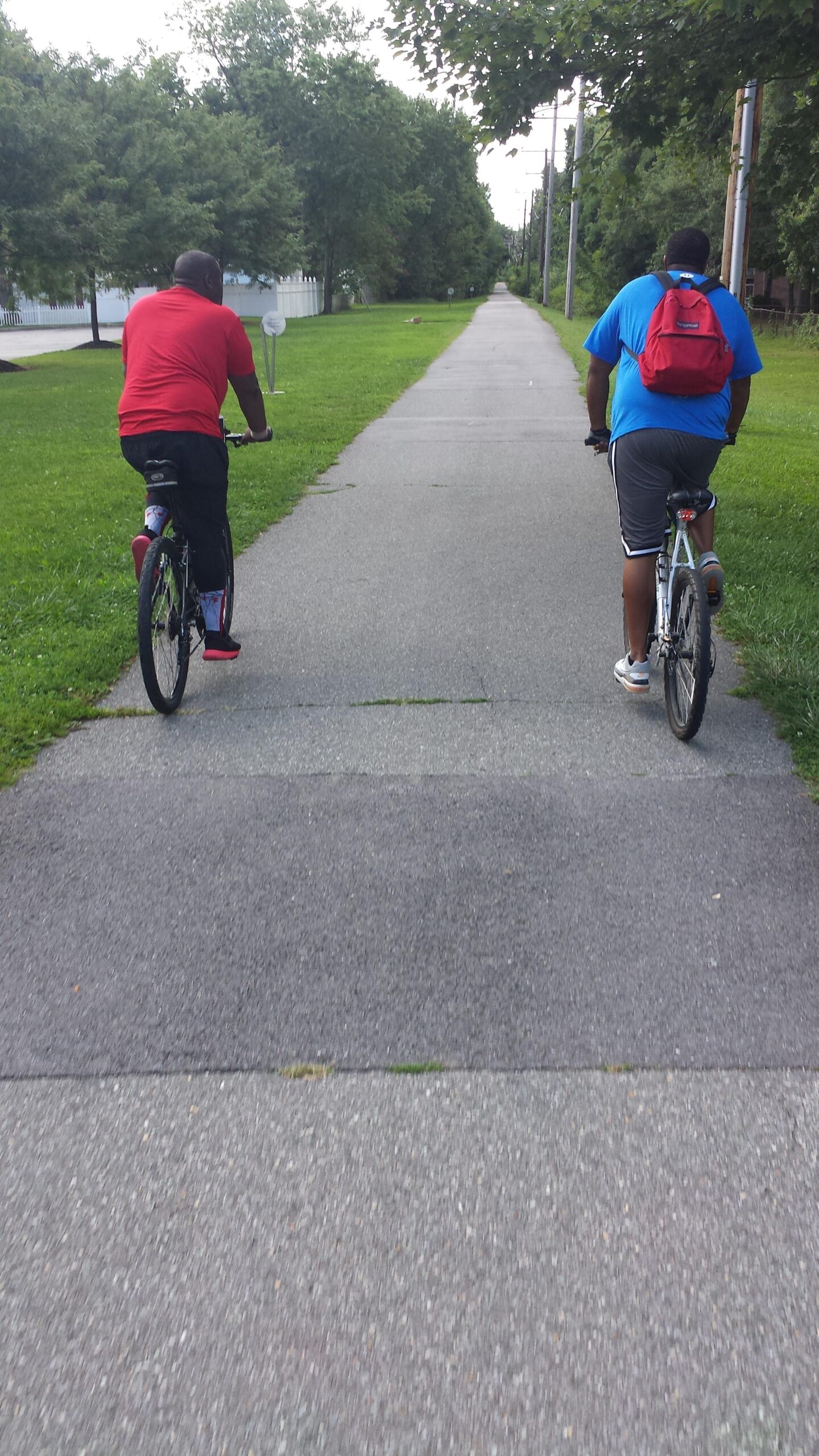 Trek 4500: Two individuals riding bicycles on a paved path surrounded by grass and trees, with a view of the path extending into the distance. One bicyclist is wearing a red shirt and the other is in a blue shirt with a backpack. The scene conveys an outdoor recreational activity on a sunny day.