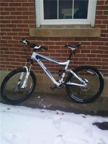 Trek Fuel EX 7: A mountain bike with a white and blue frame, resting on a snowy surface next to a brick wall with a window above it.